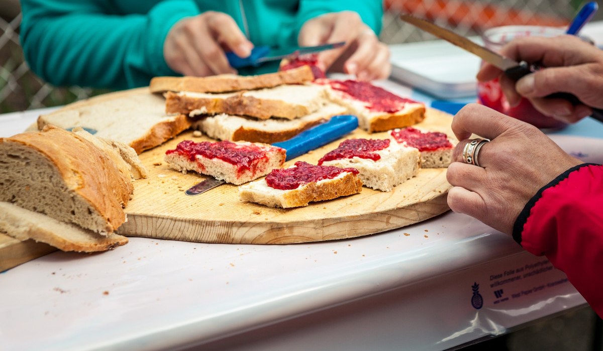 Zwei Personen schmieren Marmelade auf Brotscheiben auf einem Holzteller. Einfache Brotzeit im Freien., © hochgehberge Zwei Personen schmieren Marmelade auf Brotscheiben auf einem Holzteller. Einfache Brotzeit im Freien., © hochgehberge