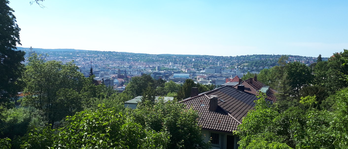 Panoramablick auf eine Stadt mit grünen Bäumen im Vordergrund und Hügeln im Hintergrund unter klarem Himmel., © SMG