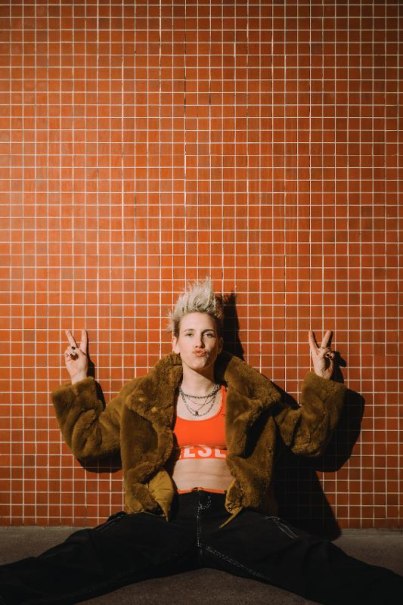 Eine Person mit blonder Frisur und brauner Jacke sitzt vor einer roten Kachelwand und zeigt das Peace-Zeichen., &copy; Music Circus Concertb&uuml;ro GmbH & Co. KG