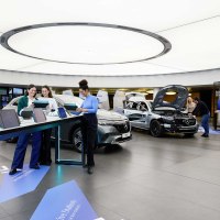 Moderner Ausstellungsraum mit Autos und interaktiven Stationen. Menschen nutzen Tablets, w&auml;hrend andere ein Auto mit ge&ouml;ffneter Motorhaube betrachten., &copy; Mercedes-Benz AG
