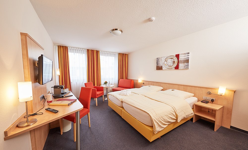 Ein modernes Hotelzimmer mit Doppelbett, Schreibtisch, Fernseher und Sitzbereich. Die Einrichtung ist in warmen Farben gehalten., © TOMAS