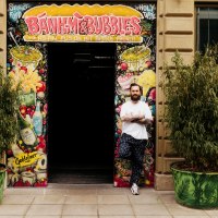 Ein Mann steht vor dem Eingang von 'Banh Mi & Bubbles', umgeben von bunten Graffiti und Pflanzen., © Banh Mi & Bubbles Restaurant / Bar