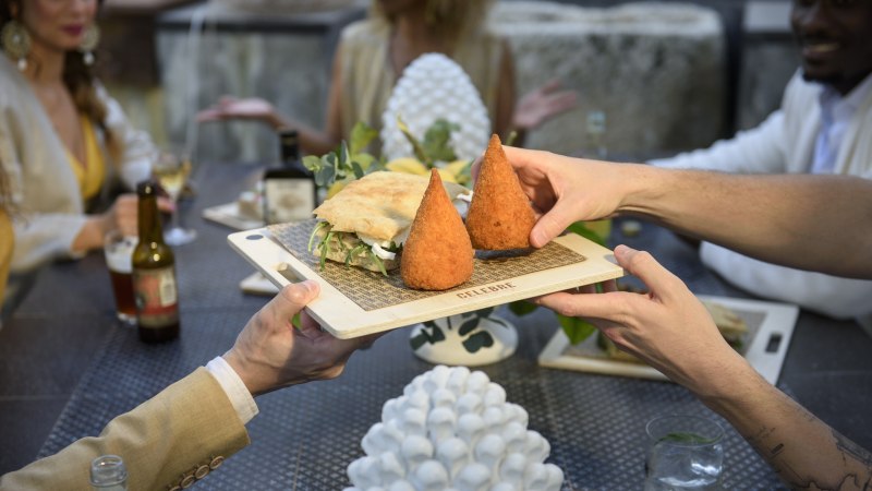 Zwei Personen reichen ein Holzbrett mit Arancini und einem Sandwich an einem Tisch mit mehreren G&auml;sten., &copy; CELEBRE