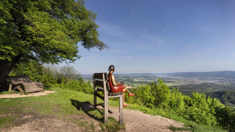 Eine Skulptur einer Frau sitzt auf einer übergroßen Bank und blickt auf eine weite, grüne Landschaft unter blauem Himmel., © Stuttgart-Marketing GmbH, Martina Denker Eine Skulptur einer Frau sitzt auf einer übergroßen Bank und blickt auf eine weite, grüne Landschaft unter blauem Himmel., © Stuttgart-Marketing GmbH, Martina Denker