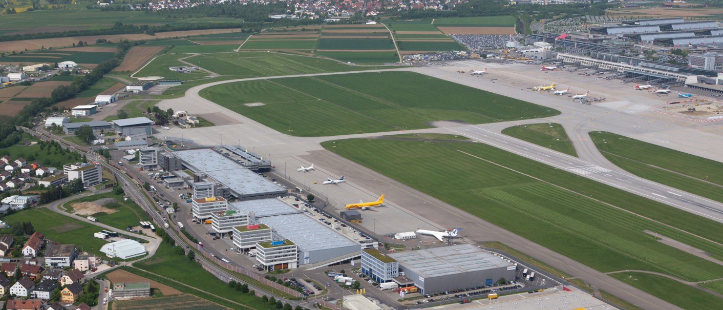 Luftaufnahme des Flughafens Stuttgart mit Landebahnen, geparkten Flugzeugen und umliegenden Geb&auml;uden. Im Hintergrund sind Felder und Wohngebiete zu sehen., &copy; SMG, Achim Mende