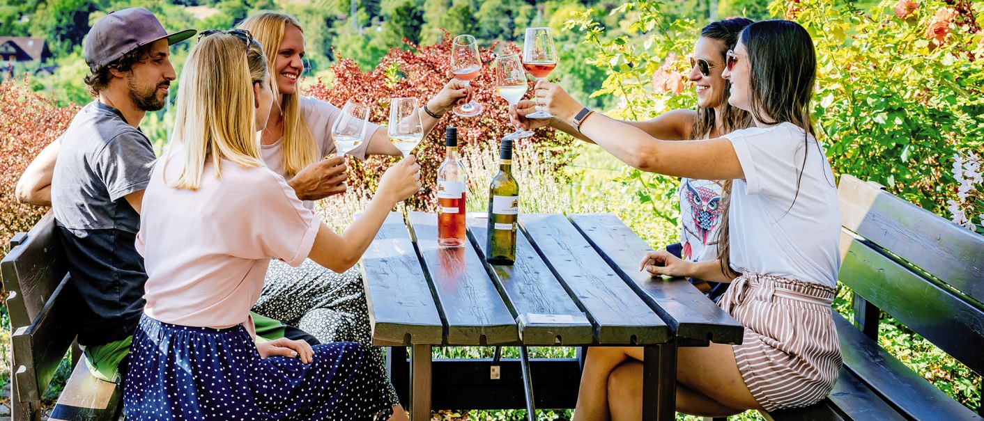 Fünf Personen stoßen mit Weingläsern an einem Holztisch im Freien an, umgeben von grüner Natur und Blumen., © SMG, Thomas Niedermüller