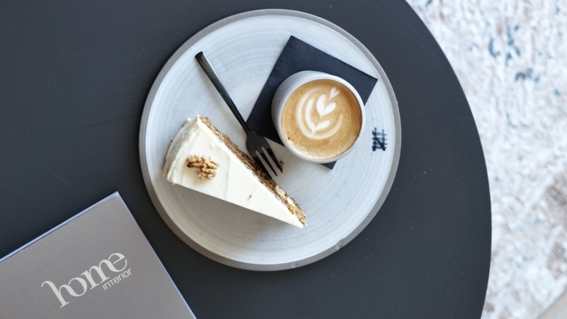 Latte mit Herz-Latte-Art und ein Stück Kuchen auf einem Teller auf schwarzem Tisch, daneben eine Karte mit „home interior“., © Taraba Latte mit Herz-Latte-Art und ein Stück Kuchen auf einem Teller auf schwarzem Tisch, daneben eine Karte mit „home interior“., © Taraba
