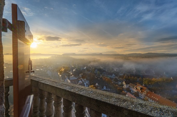Blick von einem Balkon auf eine nebelverhangene Stadt im Morgenlicht. Die Sonne geht am Horizont auf und taucht die Szene in warmes Licht., © Stadt Nürtingen Blick von einem Balkon auf eine nebelverhangene Stadt im Morgenlicht. Die Sonne geht am Horizont auf und taucht die Szene in warmes Licht., © Stadt Nürtingen