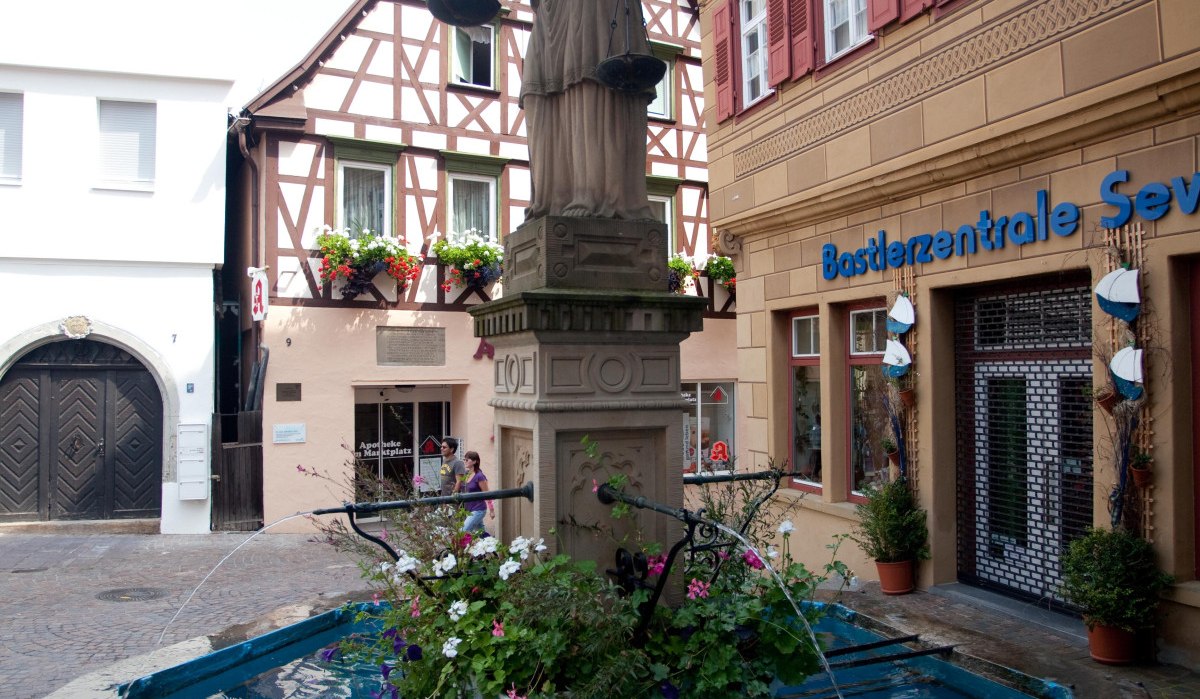 Justitia-Statue auf einem Marktbrunnen, umgeben von Fachwerkhäusern und einem Geschäft. Der Brunnen ist mit Blumen geschmückt., © Waiblingen - Stuttgart-Marketing GmbH Justitia-Statue auf einem Marktbrunnen, umgeben von Fachwerkhäusern und einem Geschäft. Der Brunnen ist mit Blumen geschmückt., © Waiblingen - Stuttgart-Marketing GmbH
