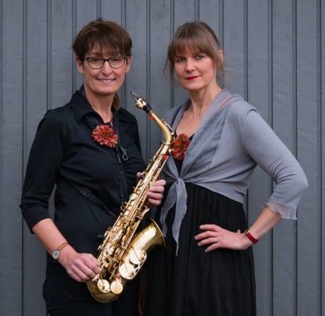 Zwei Frauen stehen vor einer grauen Wand. Eine h&auml;lt ein Saxophon, beide tragen Blumenbroschen und l&auml;cheln in die Kamera.