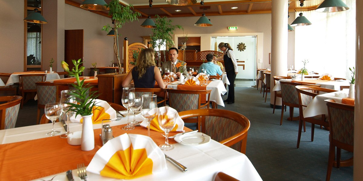 Ein elegantes Restaurant mit weiß gedeckten Tischen, orangefarbenen Servietten und Gästen, die speisen. Eine Kellnerin bedient einen Tisch., © © H-Hotels.com
