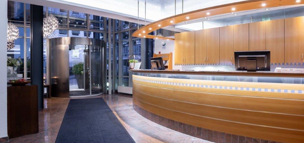 Moderne Hotelrezeption mit geschwungener Holztheke, beleuchtetem Empfangsbereich und Drehtür im Hintergrund. Elegantes, einladendes Ambiente., © Kronen Hotel GmbH Moderne Hotelrezeption mit geschwungener Holztheke, beleuchtetem Empfangsbereich und Drehtür im Hintergrund. Elegantes, einladendes Ambiente., © Kronen Hotel GmbH