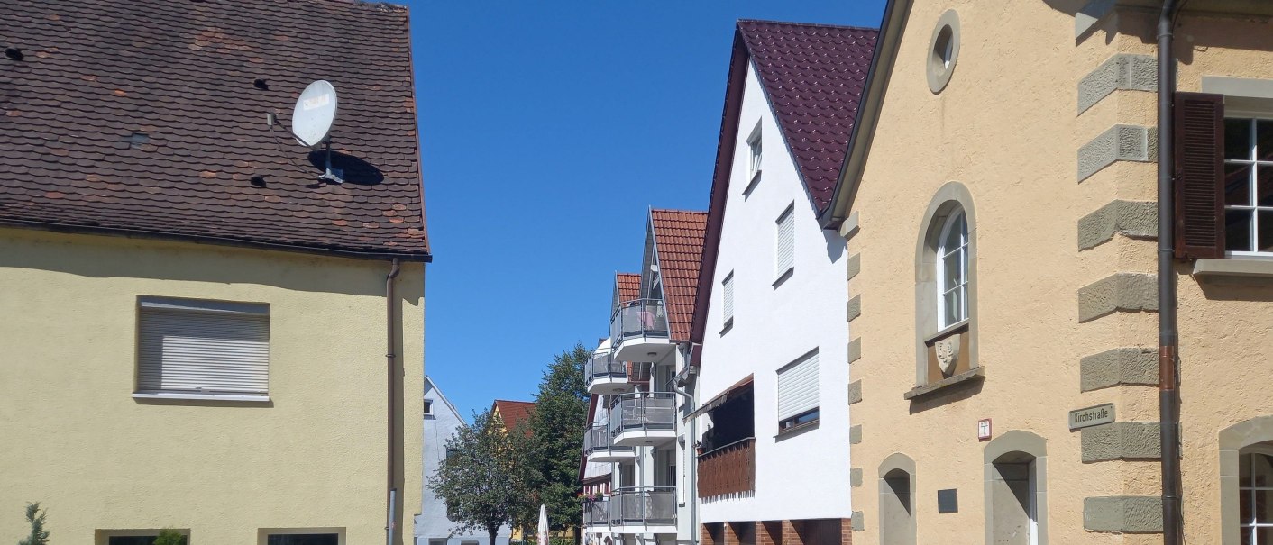 Eine ruhige Straße mit Häusern und einem klaren blauen Himmel. Die Gebäude haben rote Ziegeldächer und helle Fassaden., © Petra Natzkowski Eine ruhige Straße mit Häusern und einem klaren blauen Himmel. Die Gebäude haben rote Ziegeldächer und helle Fassaden., © Petra Natzkowski