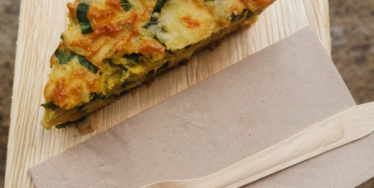 Ein Stück Quiche mit Blumen dekoriert auf einem Holzteller, daneben eine Serviette und eine Holzgabel., © n/a