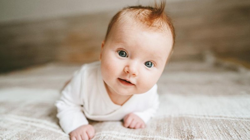 Ein Baby mit gro&szlig;en, neugierigen Augen liegt auf dem Bauch auf einer weichen Decke und blickt in die Kamera., &copy; AdobeStock_Everst