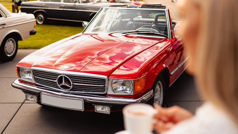 Ein roter Mercedes-Benz Oldtimer-Cabrio steht im Fokus, umgeben von weiteren klassischen Autos. Eine Person hält eine Tasse Kaffee im Vordergrund., © Mercedes-Benz AG Ein roter Mercedes-Benz Oldtimer-Cabrio steht im Fokus, umgeben von weiteren klassischen Autos. Eine Person hält eine Tasse Kaffee im Vordergrund., © Mercedes-Benz AG