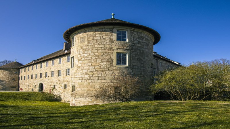 Der Turm des Schorndorfer Burgschlosses in Weitwinkelaufnahme, umgeben von grüner Wiese und Bäumen, unter einem klaren blauen Himmel., © SMG, Sarah Schmid