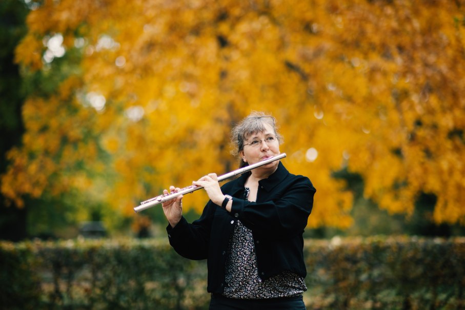 Eine Frau spielt Flöte im Freien, umgeben von herbstlich gelben Bäumen. Sie trägt eine schwarze Jacke und eine Brille., © Kathrin Beddig