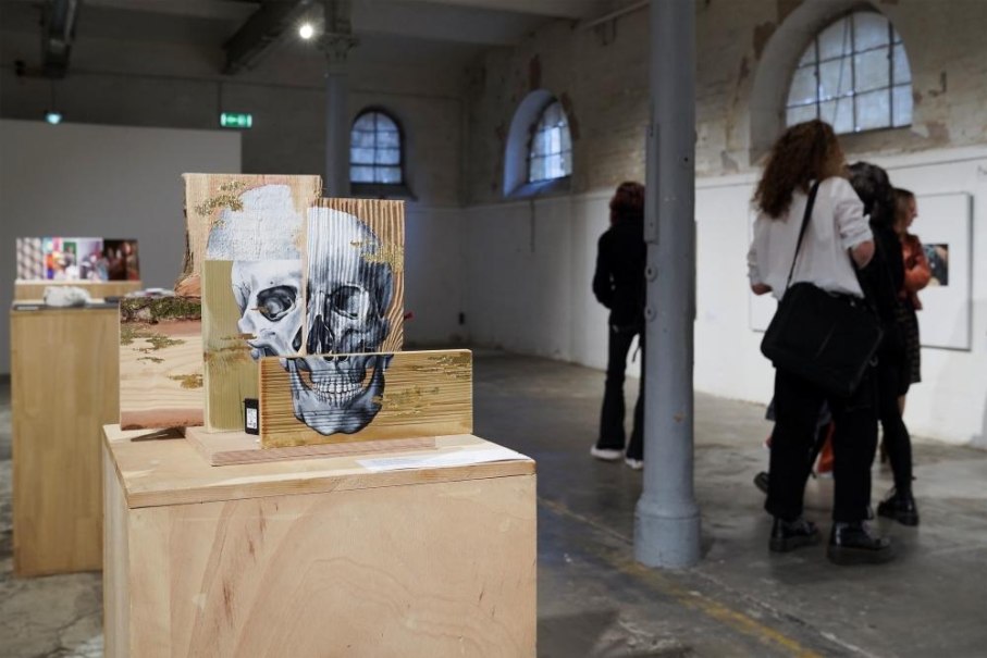 Eine Kunstinstallation mit einem Totenkopf auf Holzbl&ouml;cken in einer Galerie. Im Hintergrund sind Besucher zu sehen, die die Ausstellung betrachten., &copy; Kunstzentrum Karlskaserne