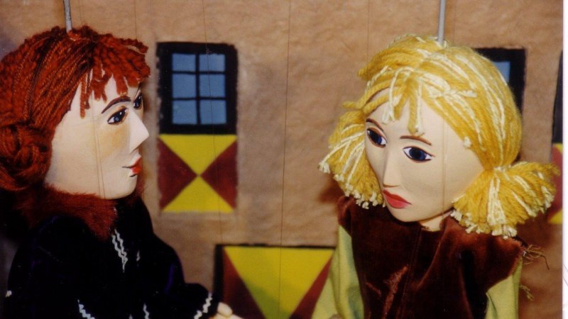 Zwei Marionetten mit roten und blonden Haaren stehen vor einem bemalten Hintergrund mit Fenstern und geometrischen Mustern., &copy; Stadtb&uuml;cherei Bad Urach