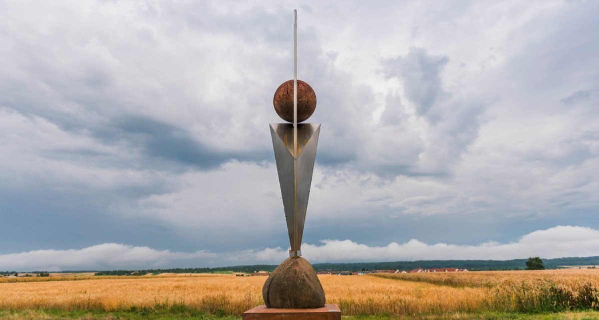 Eine moderne Skulptur mit geometrischen Formen steht auf einem Feld. Der Himmel ist bewölkt und die Landschaft ist weitläufig., © Natur.Nah. Schönbuch & Heckengäu Eine moderne Skulptur mit geometrischen Formen steht auf einem Feld. Der Himmel ist bewölkt und die Landschaft ist weitläufig., © Natur.Nah. Schönbuch & Heckengäu
