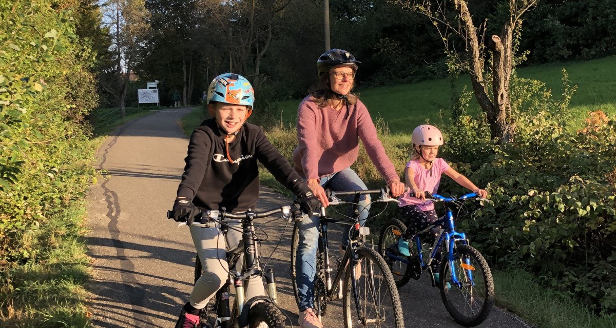 Drei Personen auf Fahrrädern fahren bei sonnigem Wetter auf einem Radweg durch eine grüne Landschaft., © T&M