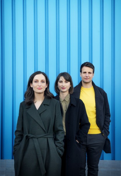 Drei Personen in dunkler Kleidung stehen vor einer blauen Wand. Die mittlere Person trägt einen gelben Pullover. Alle blicken in die Kamera., © Harald Hoffmann