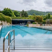 Ein Außenpool in einer Therme, umgeben von üppiger Vegetation und Hügeln im Hintergrund. Metallgeländer führen ins Wasser., © Thomas Niedermüller Ein Außenpool in einer Therme, umgeben von üppiger Vegetation und Hügeln im Hintergrund. Metallgeländer führen ins Wasser., © Thomas Niedermüller