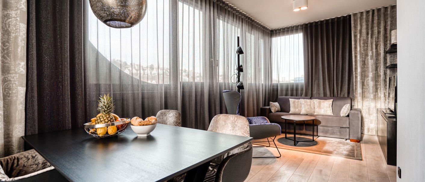 Moderne Suite mit stilvollem Esstisch, Obstschale, gemütlichem Sofa und großen Fenstern mit eleganten Vorhängen., © CLOUD N°7 APARTMENTS GmbH