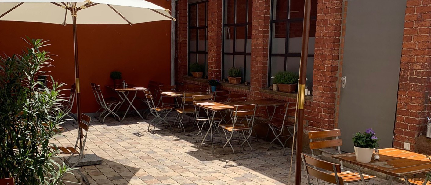 Gemütlicher Innenhof mit Holztischen und Stühlen, Sonnenschirm und Pflanzen. Rote Backsteinwände und gepflasterter Boden schaffen eine einladende Atmosphäre., © Gustav Gastro GmbH