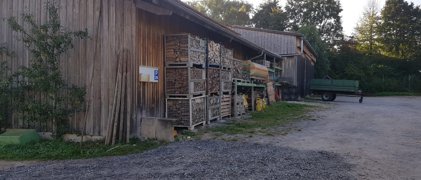 Holzhütte mit gestapeltem Holz in Metallkisten, daneben ein grüner Anhänger. Im Hintergrund Bäume und ein Parkplatzschild., © Mannschreck-Weine