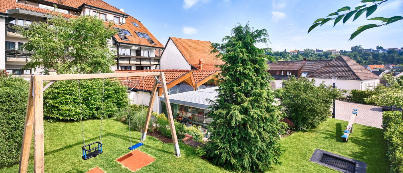 Ein grüner Garten mit Schaukel, Wippe und Trampolin, umgeben von Häusern mit roten Dächern., © Hotel Restaurant Lamm