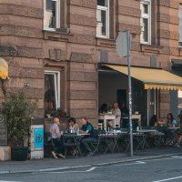 Caf&eacute; mit gelbem Vordach an einer Stra&szlig;enecke. G&auml;ste sitzen drau&szlig;en an Tischen. Schild mit 'Lumen' und Stra&szlig;enschild 'Schwabstr.' sichtbar., &copy; SMG, Sarah Schmid