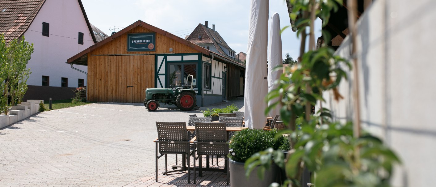 Ein rustikales Gebäude mit der Aufschrift 'BREWSCHEUNE', ein grüner Traktor davor. Im Vordergrund ein Sitzbereich mit Tischen und Stühlen., © SDV Werbestudio