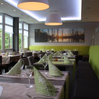 Modernes Restaurant mit gedeckten Tischen, gr&uuml;nen Servietten und gro&szlig;en Fenstern. An der Wand h&auml;ngt ein Bild mit Sonnenuntergangsmotiv., &copy; Hotel am Schlossberg Herrenberg