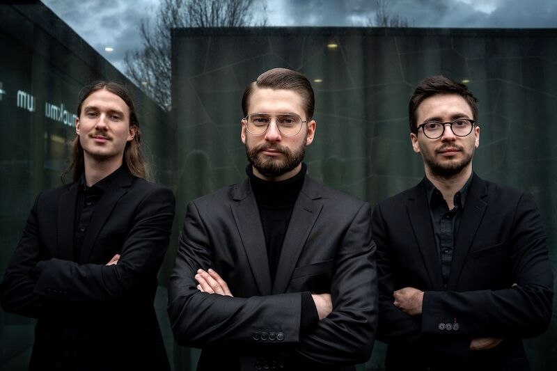 Drei M&auml;nner in schwarzen Anz&uuml;gen posieren selbstbewusst vor einer modernen Glasfassade. Der Himmel ist bew&ouml;lkt, was der Szene eine dramatische Note verleiht., &copy; Interessengemeinschaft Kultur Sindelfingen / B&ouml;blingen e.V.