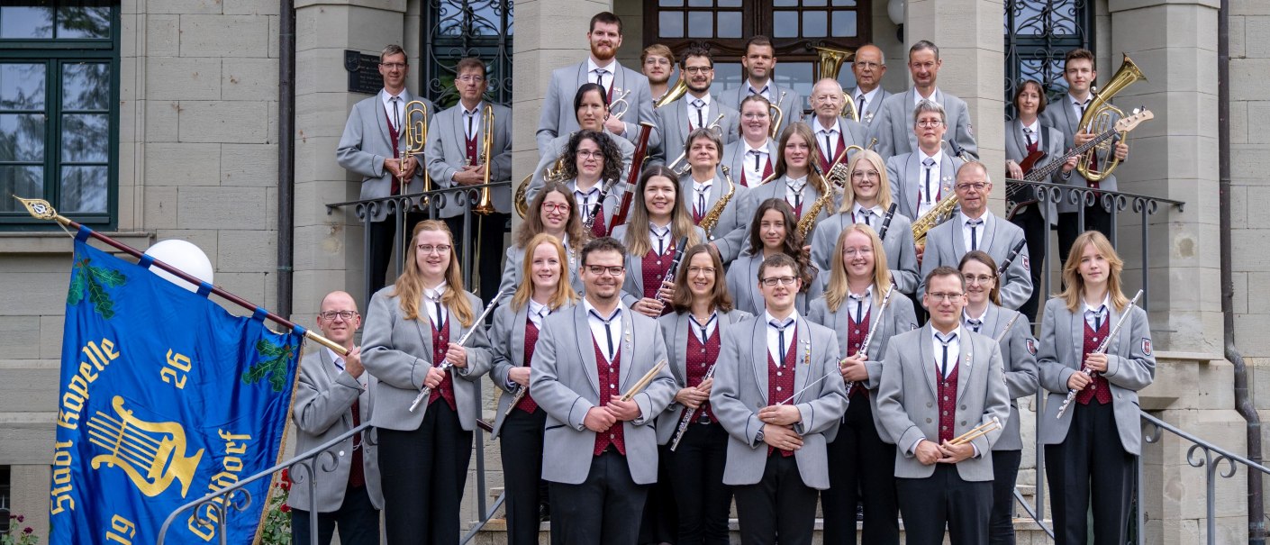 Gruppenfoto der Musiker der Stadtkapelle mit dunkelroter Weste, grauem Jacket und schwarzen Hosen. Die Musiker stehen auf den Treppenstufen vor dem Eingang des Neuen Schlosses und halten Musikinsturmente in den Händen, © Stadtkapelle Gaildorf