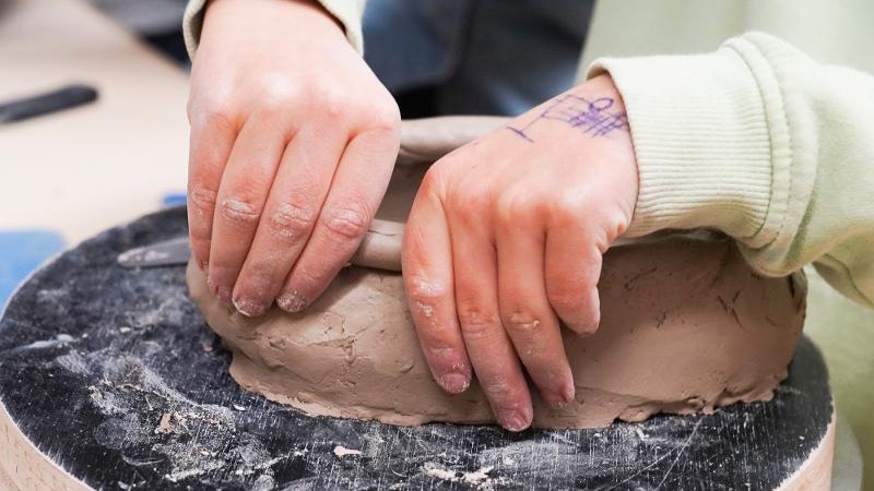 Ein Kind formt Ton mit den Händen auf einem Drehteller. Die Hände sind leicht mit Ton bedeckt, und auf der Hand ist eine Zeichnung zu sehen., © Kunstschule Labyrinth Ein Kind formt Ton mit den Händen auf einem Drehteller. Die Hände sind leicht mit Ton bedeckt, und auf der Hand ist eine Zeichnung zu sehen., © Kunstschule Labyrinth