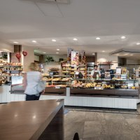 Eine Bäckerei mit Theke voller Backwaren. Zwei Mitarbeiter bedienen Kunden. Die Einrichtung ist modern und einladend., © Trölsch GmbH Eine Bäckerei mit Theke voller Backwaren. Zwei Mitarbeiter bedienen Kunden. Die Einrichtung ist modern und einladend., © Trölsch GmbH