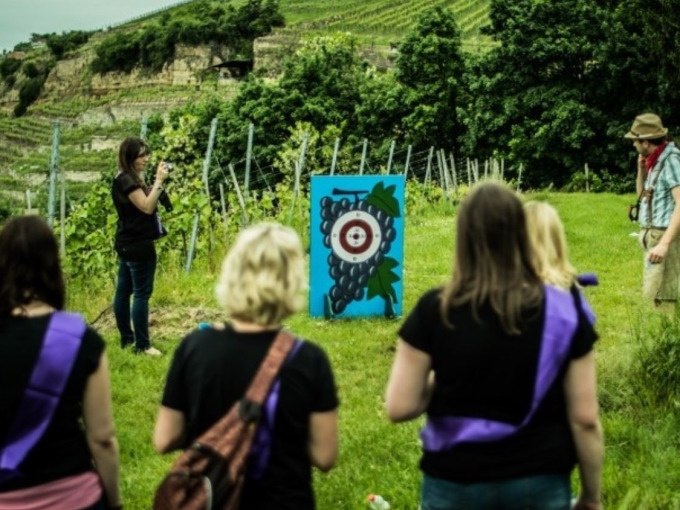 Menschen in einem Weinberg bei einer Weinsafari, mit einer Zielscheibe in Form einer Traube. Im Hintergrund sind Weinreben und Hügel zu sehen., © Cool-Tours StattReisen Menschen in einem Weinberg bei einer Weinsafari, mit einer Zielscheibe in Form einer Traube. Im Hintergrund sind Weinreben und Hügel zu sehen., © Cool-Tours StattReisen