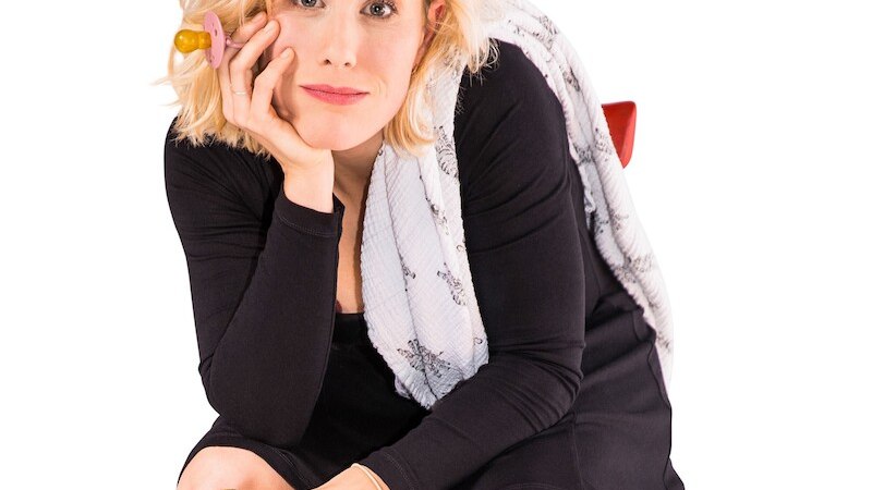 Eine Frau mit blonden Haaren sitzt in einem schwarzen Kleid, h&auml;lt einen Schnuller am Ohr und einen goldenen Becher in der Hand., &copy; Mauerwerk GmbH