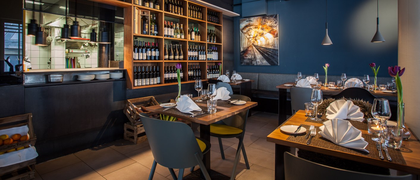 Stilvolles Restaurant mit gedeckten Tischen, Weinregalen und moderner Beleuchtung. Eine offene Küche und ein Kunstwerk an der Wand sind sichtbar., © Belser