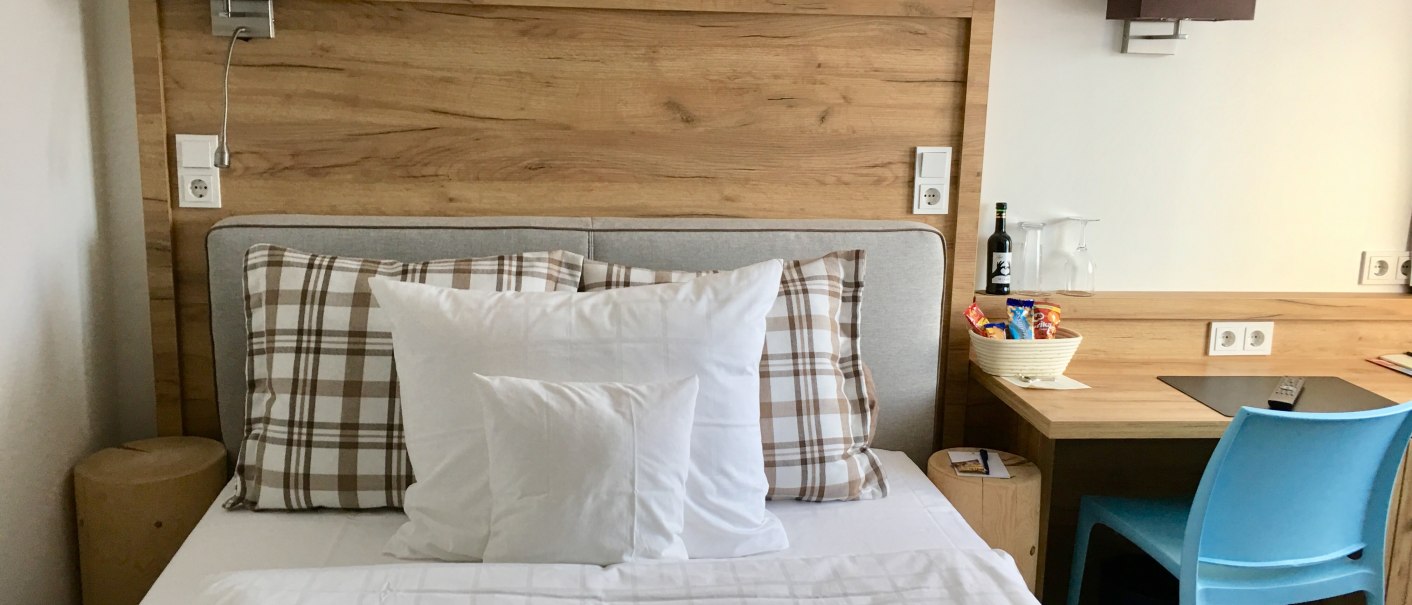 Modernes Hotelzimmer mit Einzelbett, Holzkopfteil, Schreibtisch, blauem Stuhl und Snacks auf dem Tisch., © TOMAS Modernes Hotelzimmer mit Einzelbett, Holzkopfteil, Schreibtisch, blauem Stuhl und Snacks auf dem Tisch., © TOMAS