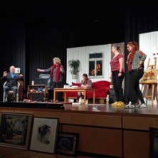 Schauspieler auf einer B&uuml;hne in einem Wohnzimmer-Set. Im Vordergrund sind Gem&auml;lde zu sehen. Die Szene wirkt lebhaft und dynamisch., &copy; links im Bild