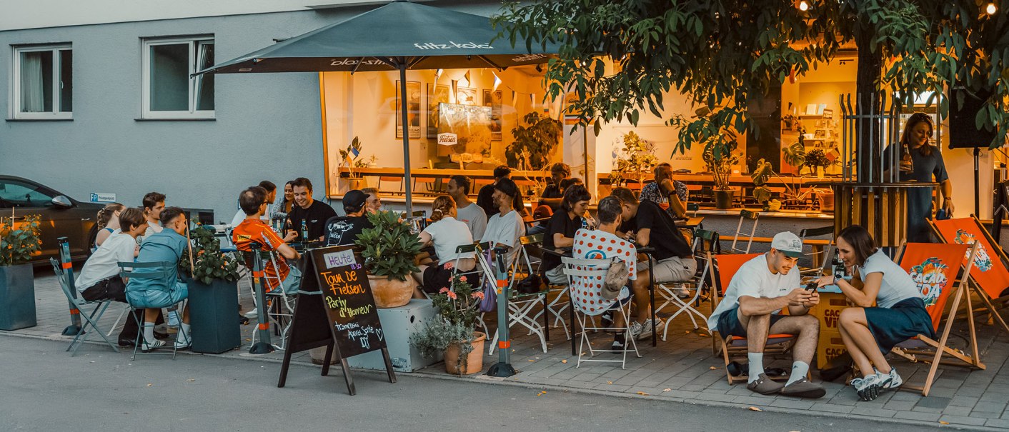 Abendliche Szene auf einer Terrasse mit vielen Menschen, die unter einem Sonnenschirm sitzen und sich unterhalten. Im Hintergrund leuchtet ein Caf&eacute;., &copy; Radcaf&eacute; Fietsen