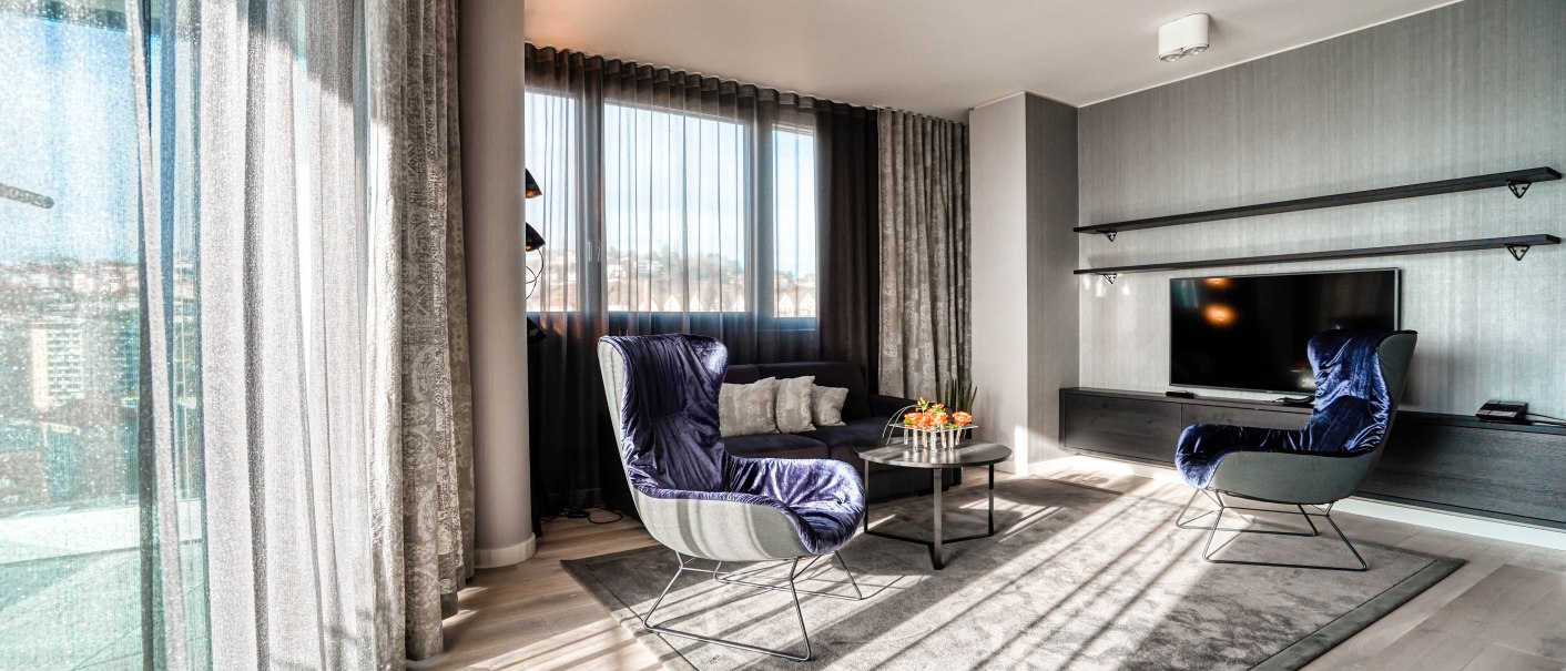 Moderne Suite mit großen Fenstern, blauen Sesseln, Flachbildfernseher und Couchtisch auf Teppich., © CLOUD N°7 APARTMENTS GmbH