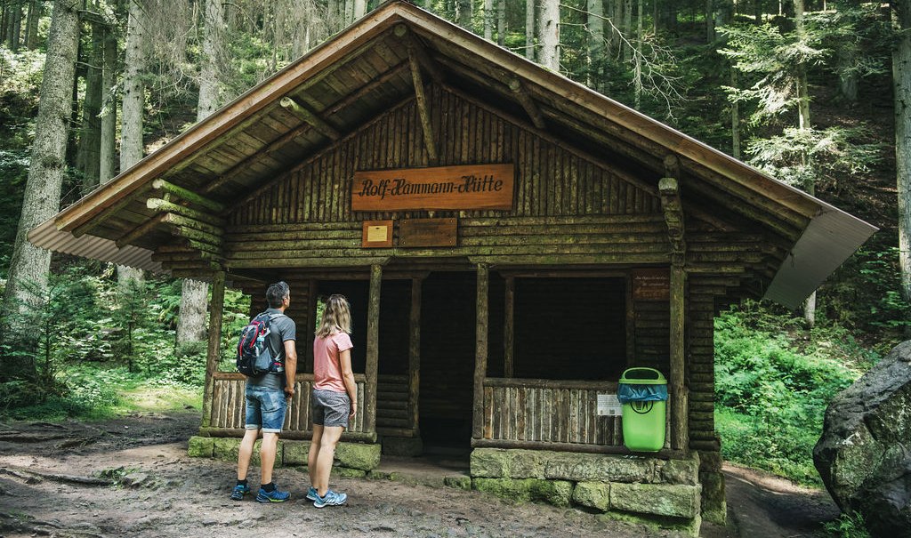 Zwei Personen betrachten die Rolf Hamann Hütte im Wald. Die Hütte ist aus Holz und von Bäumen umgeben., © LOCHER Fotodesign&Manufaktur Zwei Personen betrachten die Rolf Hamann Hütte im Wald. Die Hütte ist aus Holz und von Bäumen umgeben., © LOCHER Fotodesign&Manufaktur