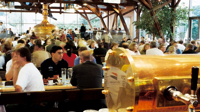 Belebtes Brauhaus mit vielen Menschen an Tischen, Kupferkessel im Hintergrund, Holzbalken und große Fenster., © SMG Belebtes Brauhaus mit vielen Menschen an Tischen, Kupferkessel im Hintergrund, Holzbalken und große Fenster., © SMG