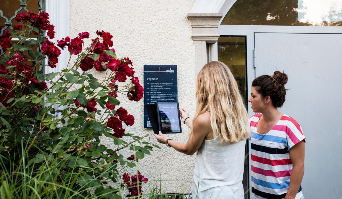 Zwei Frauen stehen vor einer Tafel am Vogthaus. Eine scannt mit einem Tablet einen QR-Code. Rote Rosen blühen daneben., © Stadt Kirchheim unter Teck