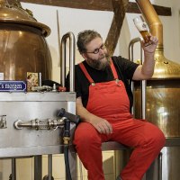 Ein Mann in roter Latzhose sitzt auf einer Kupferbrauanlage und betrachtet ein Bierglas. Im Hintergrund sind Gläser und Brauzubehör zu sehen., © Torsten Wenzler